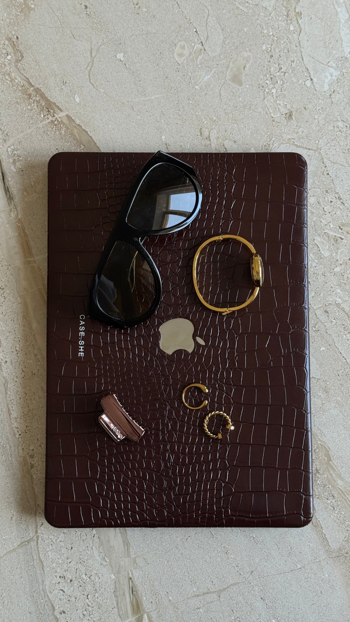 MacBook у чохлі з фактурою під шкіру крокодила та написом CASE.SHE, на ньому окуляри, браслет і прикраси