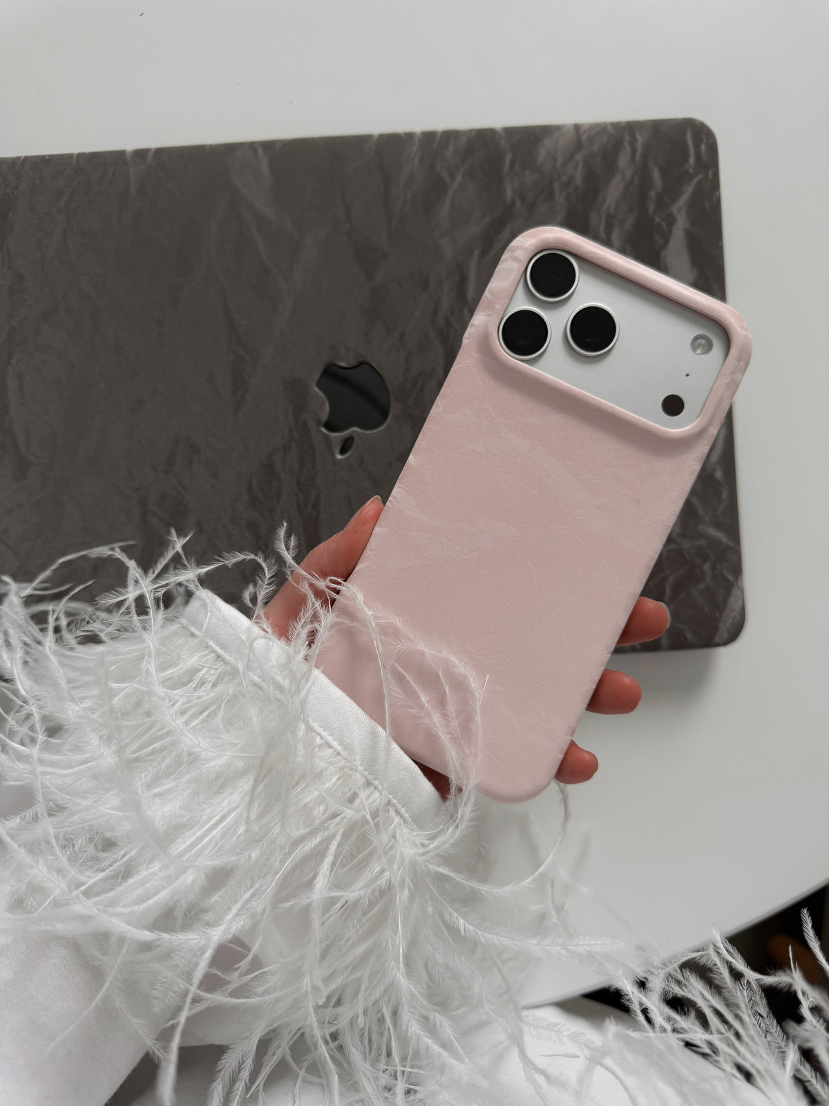 Рожевий чохол «Оксамит» на iPhone у руці з пір’ям на рукаві на тлі сірого чохла для MacBook