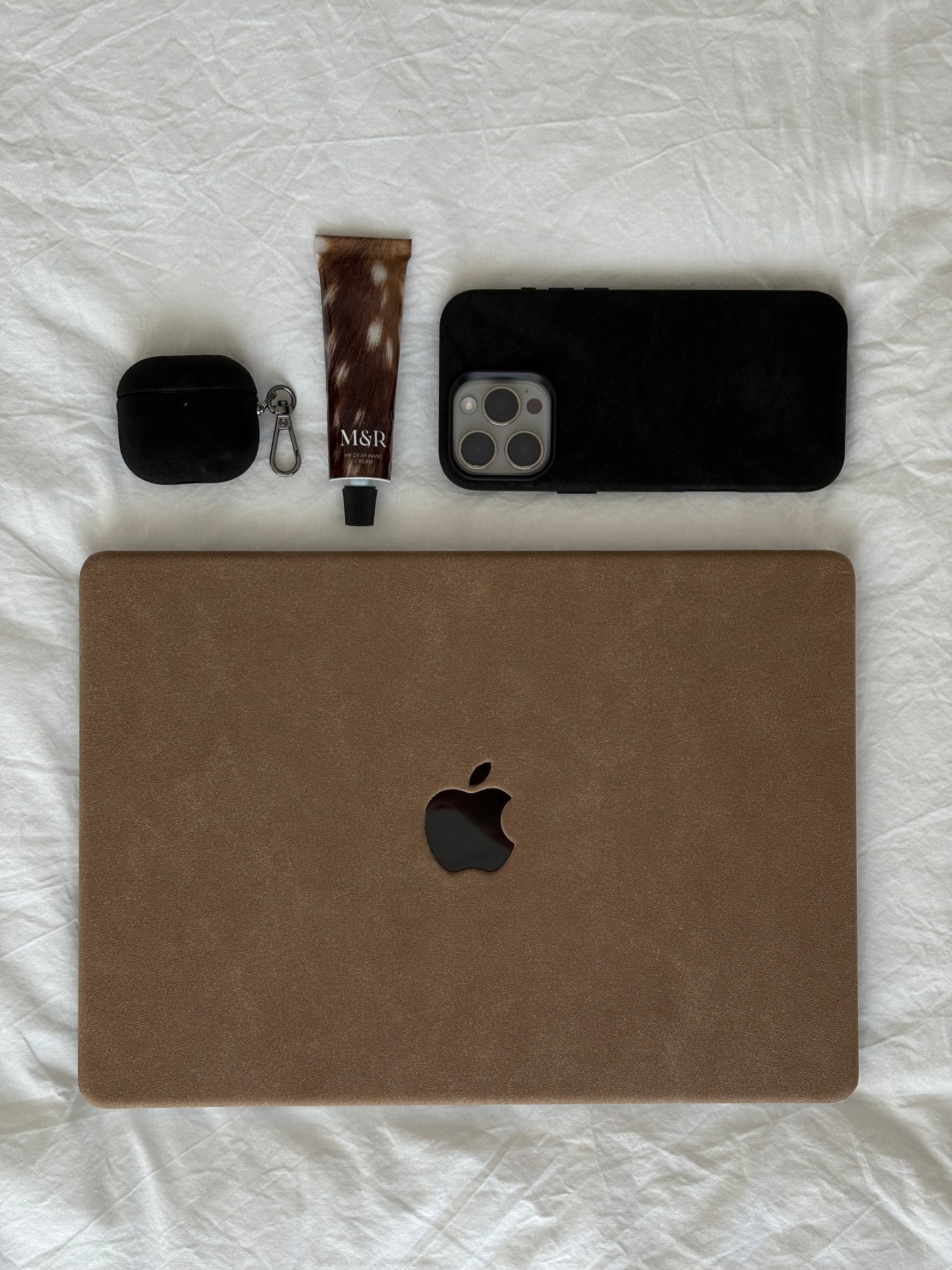 Коричневий чохол «Замшева м’якість» для MacBook на білій тканині поруч з iPhone і навушниками в чорних чохлах