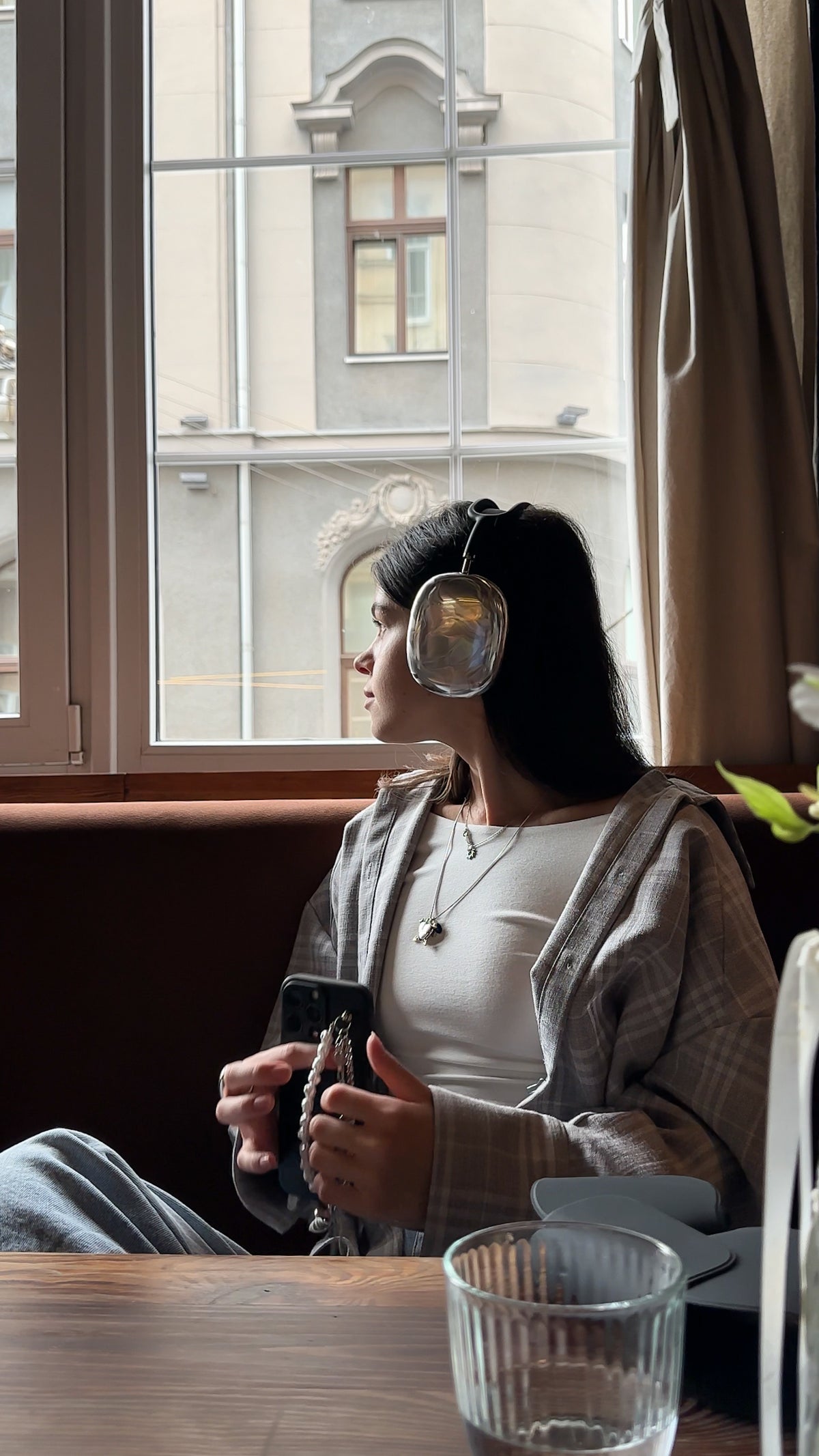 Жінка в навушниках AirPods Max у сріблястих накладках сидить біля вікна в кав’ярні