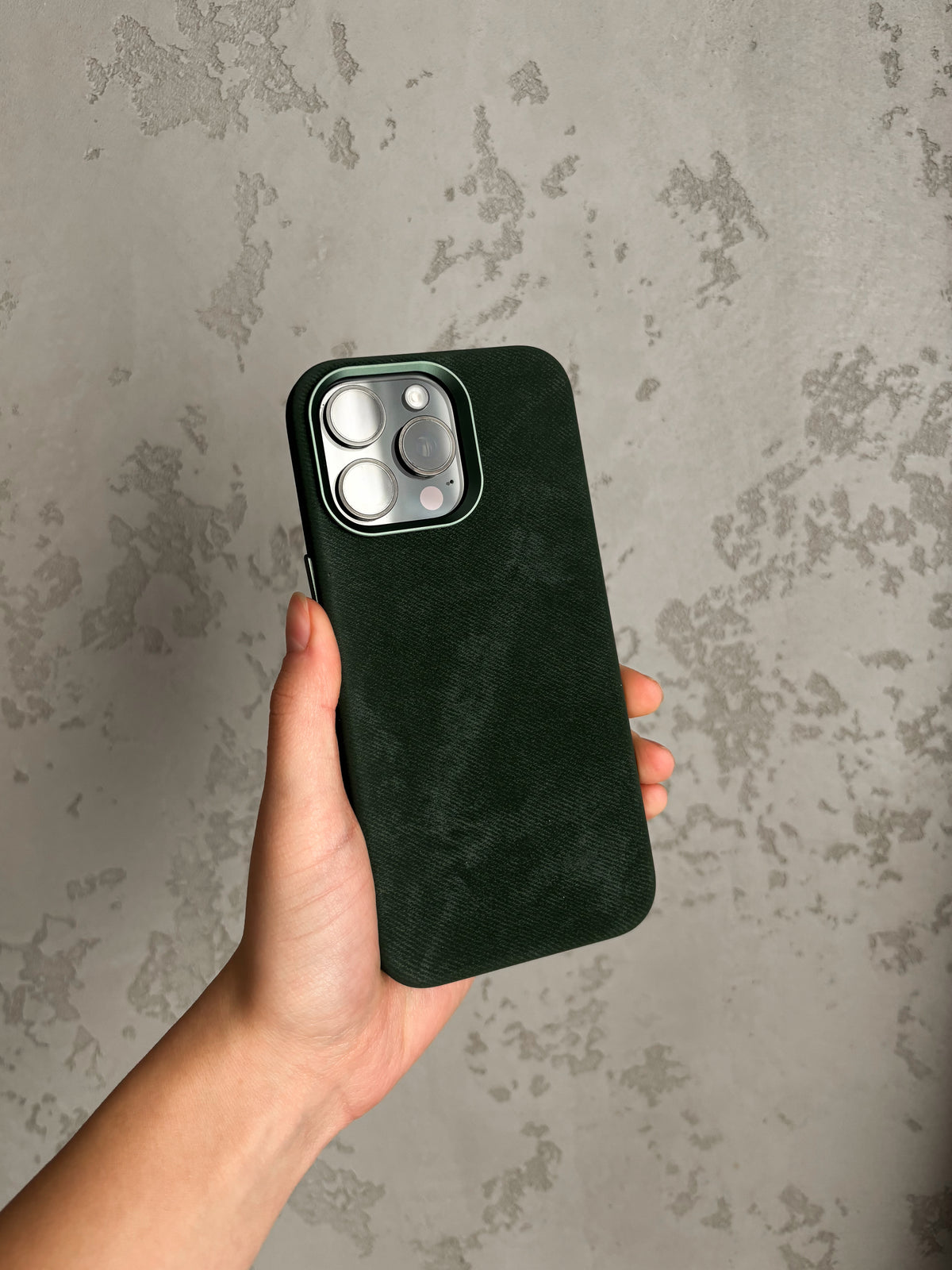 Темно-зелений чохол «Замшева м’якість» на iPhone у руці на тлі фактурної стіни